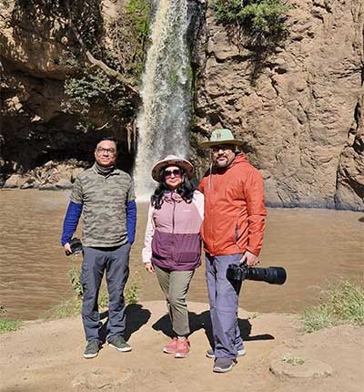 Family safari in Kenya with three travelers posing in front of a scenic waterfall surrounded by rocky cliffs during an unforgettable African travel adventure.